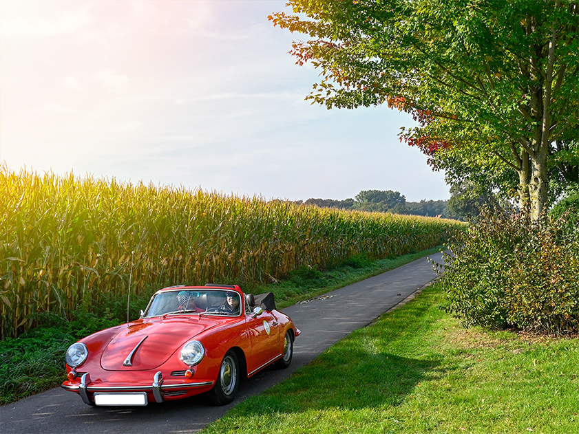Bild eines Oldtimers im Sonnenschein auf einem Feldweg; Ein historischer Porsche fährt neben einem Maisfeld; ADAC Bad Sassendorf Classic