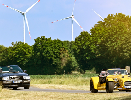 Ein gelbes und ein schwarzes Auto in einer sonnigen Landschaft mit zwei Windrädern im Hintergrund
